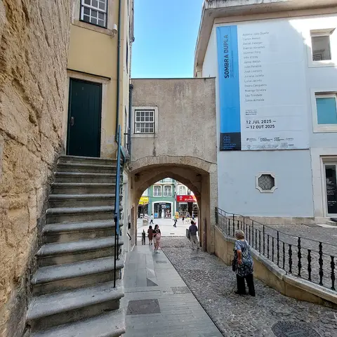 Arco de Almedina em Coimbra, com escadaria de pedra e cartaz de exposição na parede branca.
