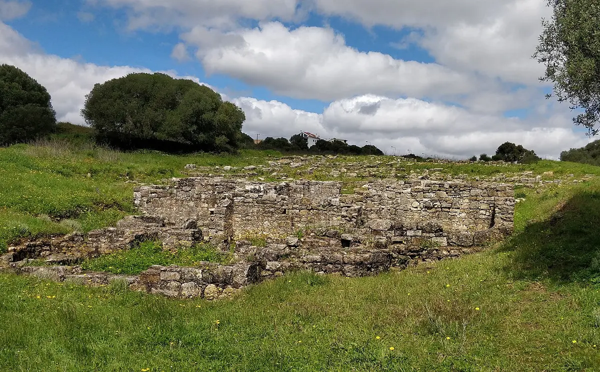 Ruínas de muros de pedra da Vila Romana de Freiria rodeadas de relva verde e céu com nuvens