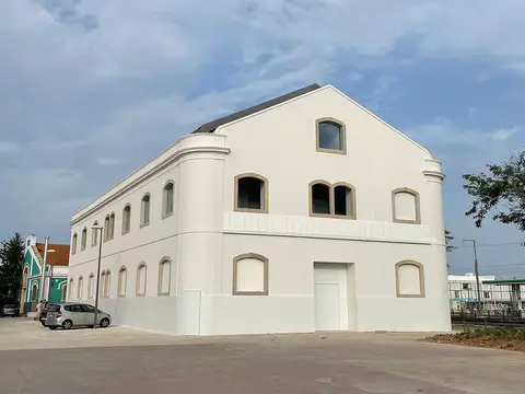 Edifício branco de dois pisos com janelas em arco, sob céu nublado, e carros estacionados ao lado.