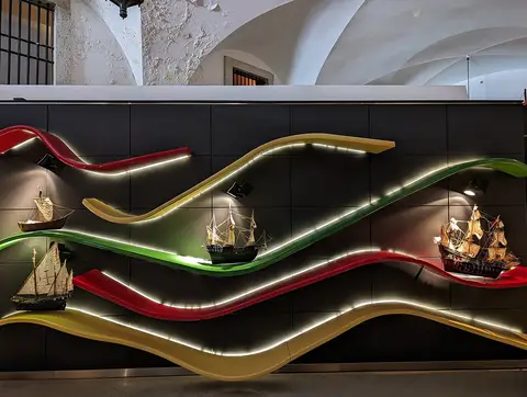 Parede expositiva com modelos de barcos antigos sobre ondas coloridas iluminadas, no Lisboa Story Centre