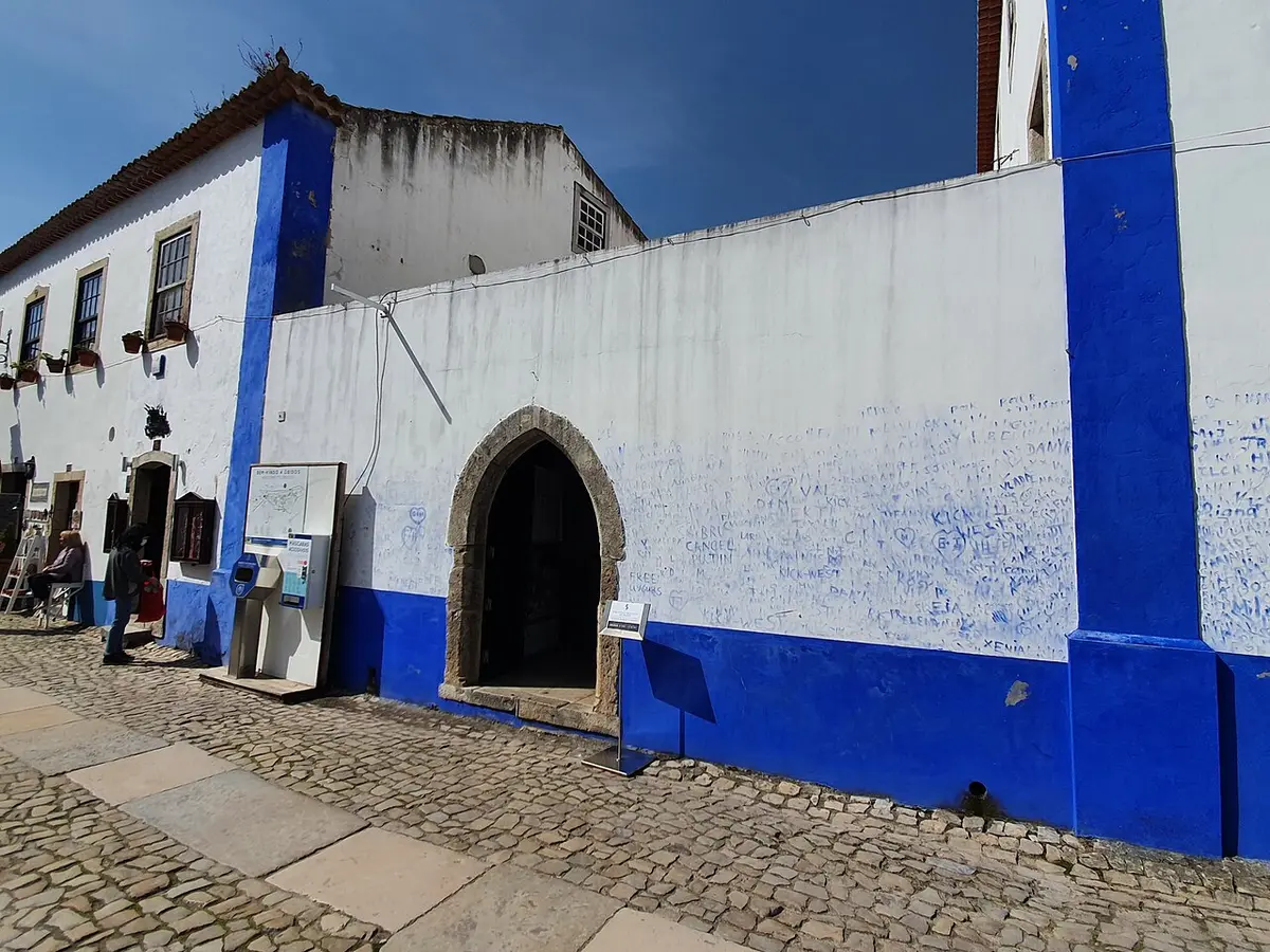 Fachada branca e azul da Biblioteca Municipal de Óbidos com porta em arco e rua de calçada