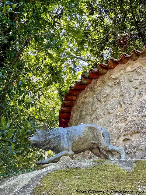 Escultura de lobo em pedra junto a muro de granito com telhado de telha, rodeada de árvores