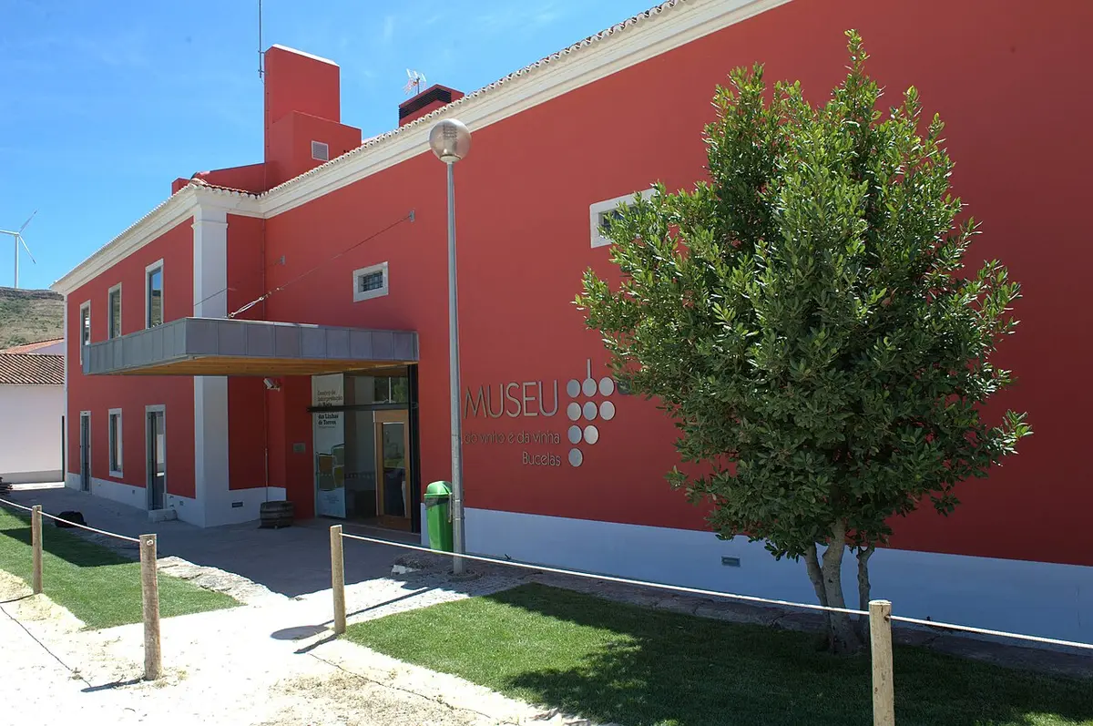 Fachada vermelha do Museu do Vinho e da Vinha de Bucelas, com entrada e árvore em frente.
