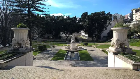 Jardim com grandes vasos de pedra sobre leões, relvado e repuxo central, rodeado de árvores.