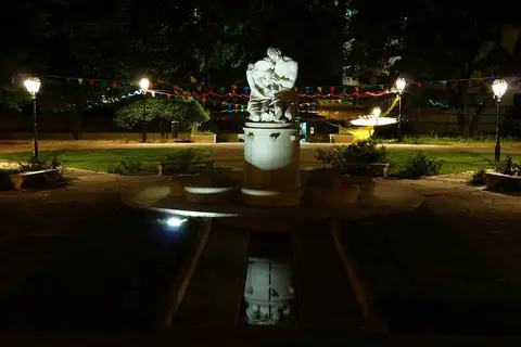 Fonte com escultura iluminada à noite num jardim com bandeirinhas coloridas e candeeiros