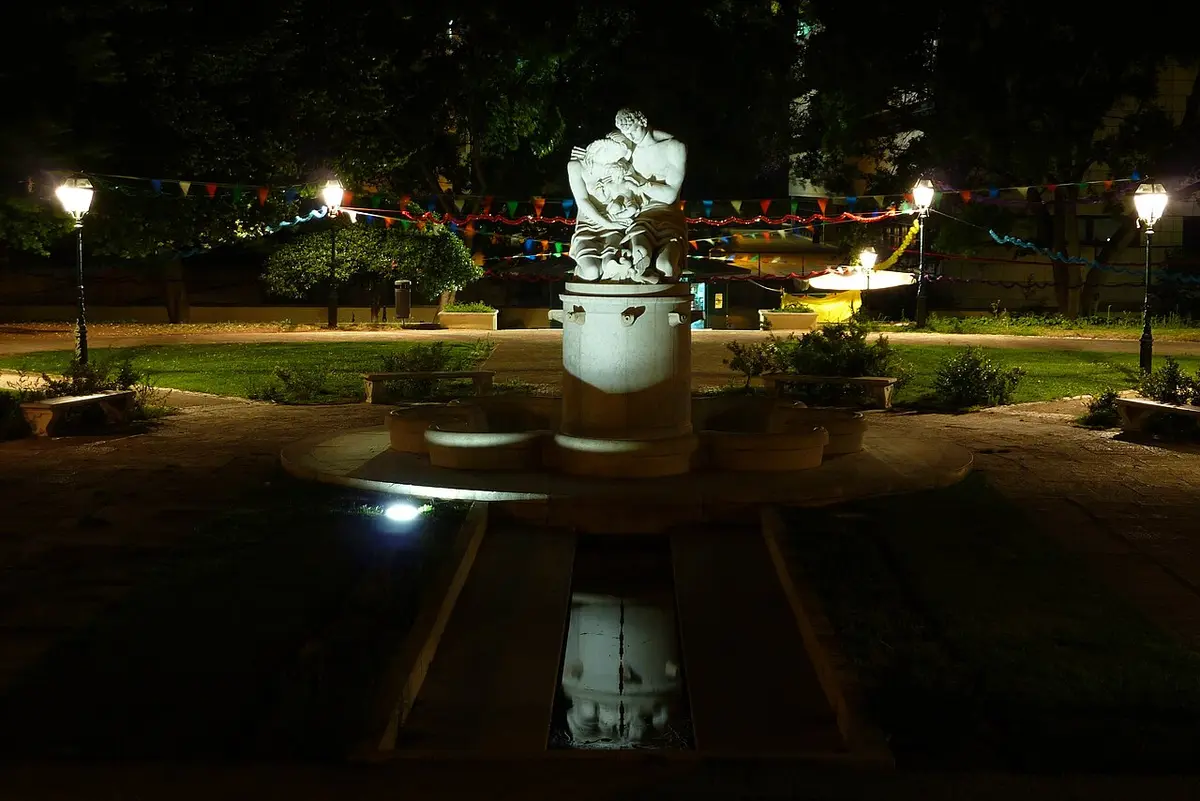 Fonte com escultura iluminada à noite num jardim com bandeirinhas coloridas e candeeiros