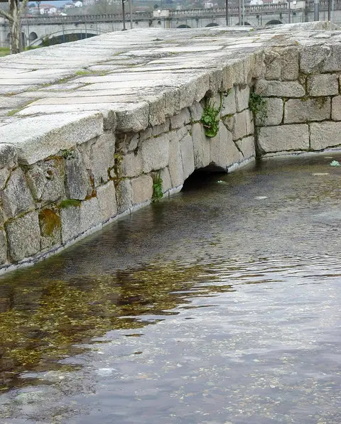 Estrutura de granito junto à água, nas Termas Romanas de Chaves, com a ponte romana ao fundo.