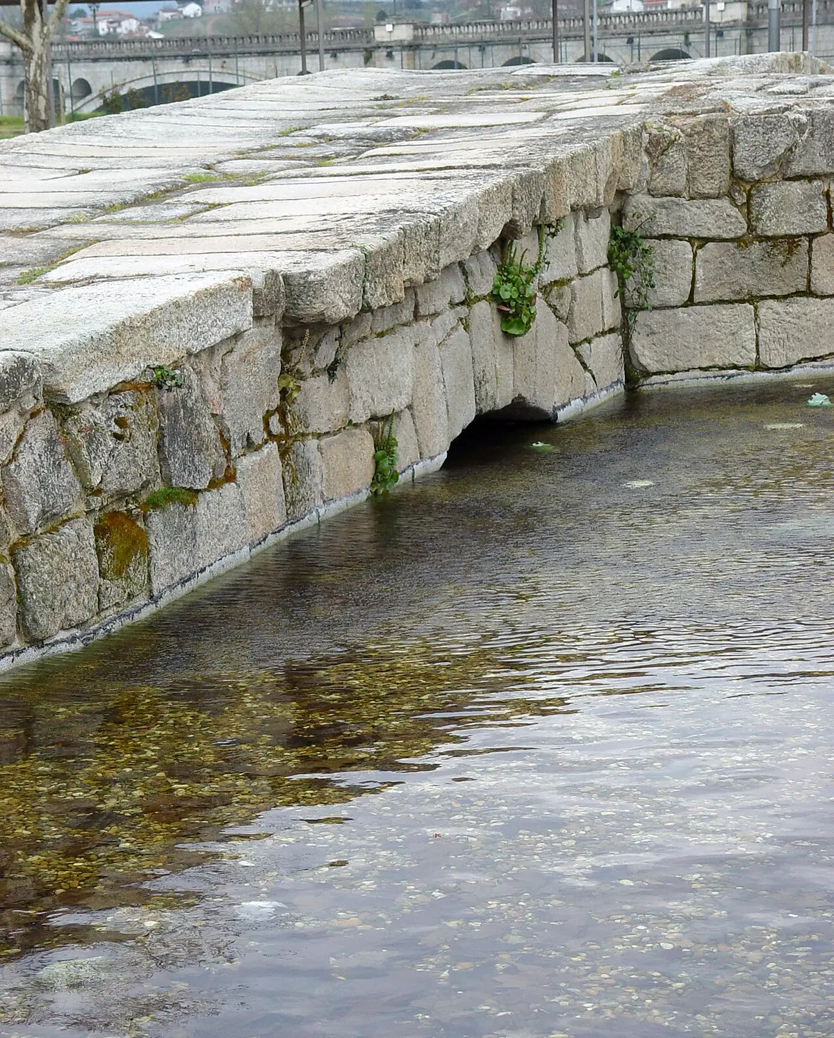 Estrutura de granito junto à água, nas Termas Romanas de Chaves, com a ponte romana ao fundo.