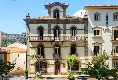 Edifício histórico de três pisos com varandas de ferro e janelas em arco, num largo ensolarado