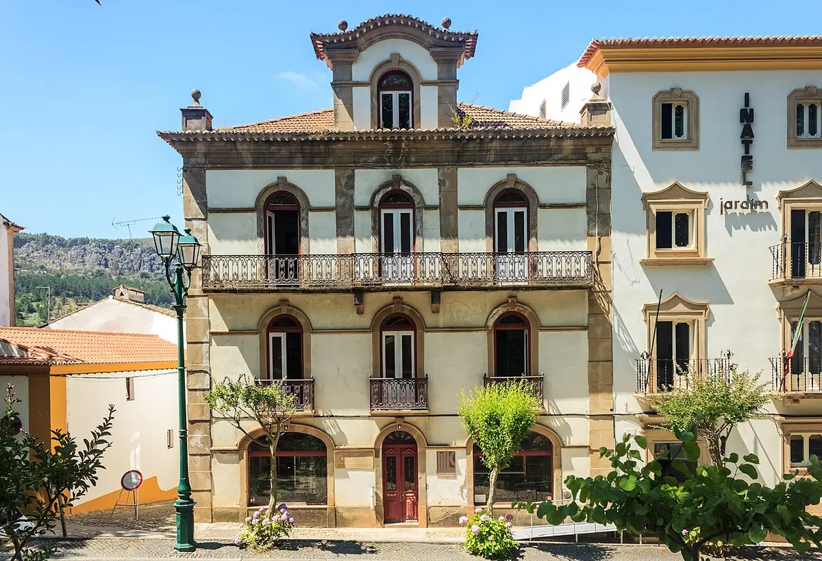 Edifício histórico de três pisos com varandas de ferro e janelas em arco, num largo ensolarado