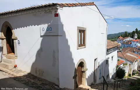 Edifício caiado da Sinagoga de Castelo de Vide, com placa do museu, em rua de calçada em declive.