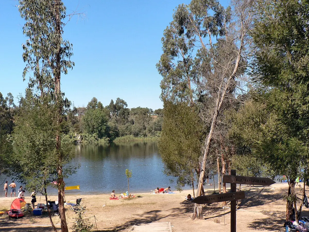 Praia fluvial do Açude do Gameiro com areal, eucaliptos e placa indicativa junto à água