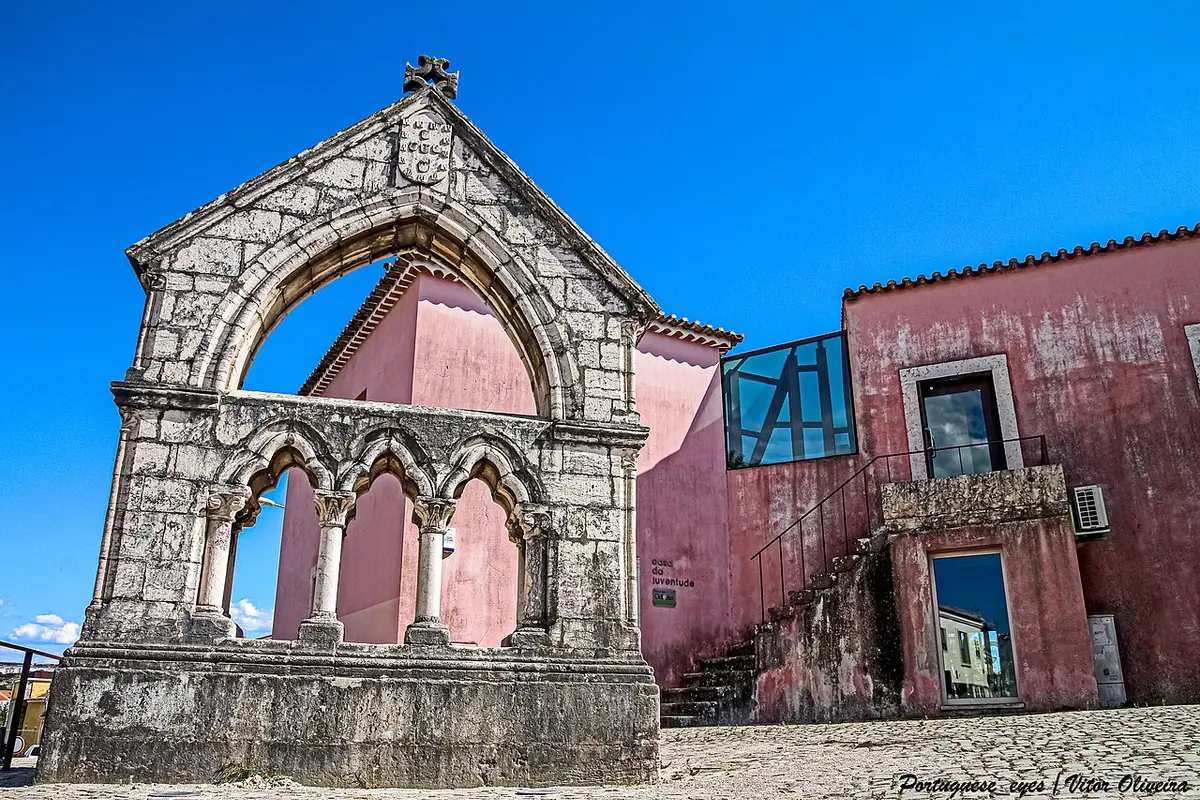 Memorial gótico em pedra junto a edifício rosa da Casa da Juventude, em Odivelas, sob céu azul
