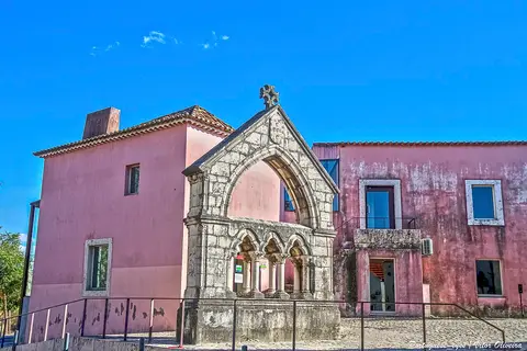 Edifício rosa com memorial gótico em pedra à frente, sob céu azul, em Odivelas