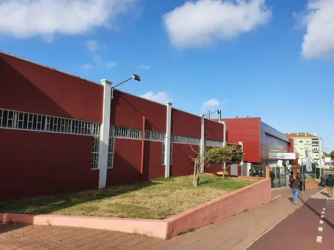 Exterior do edifício vermelho da Piscina Municipal de Ouressa, com relvado e passeio em dia de sol.
