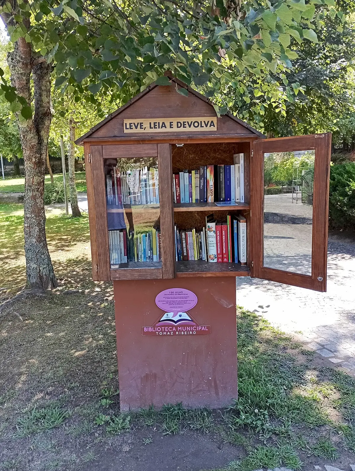 Armário de troca de livros em madeira num parque, com a inscrição «Leve, Leia e Devolva» e logótipo da Biblioteca Municipal Tomaz Ribeiro