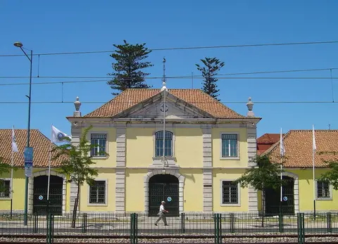Fachada amarela da Cordoaria Nacional em Lisboa, com bandeiras e linhas de comboio em primeiro plano