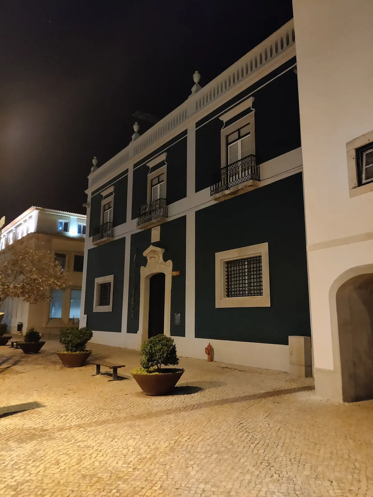 Fachada verde da Biblioteca Pública Municipal de Setúbal iluminada à noite, com praça calcetada e vasos.