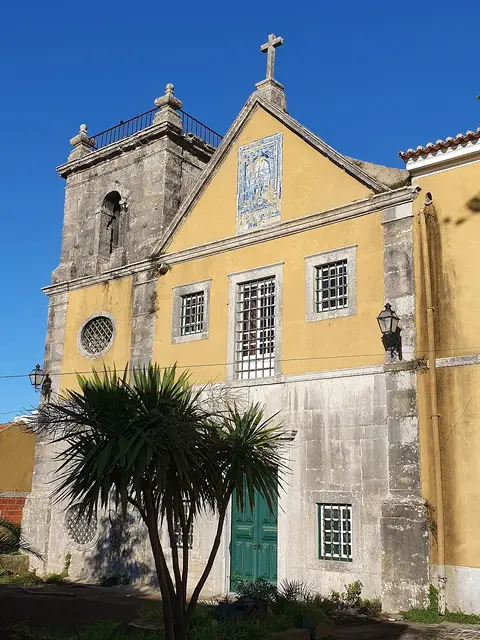 Fachada amarela de igreja com painel de azulejos, cruz e torre sineira, junto a uma palmeira.