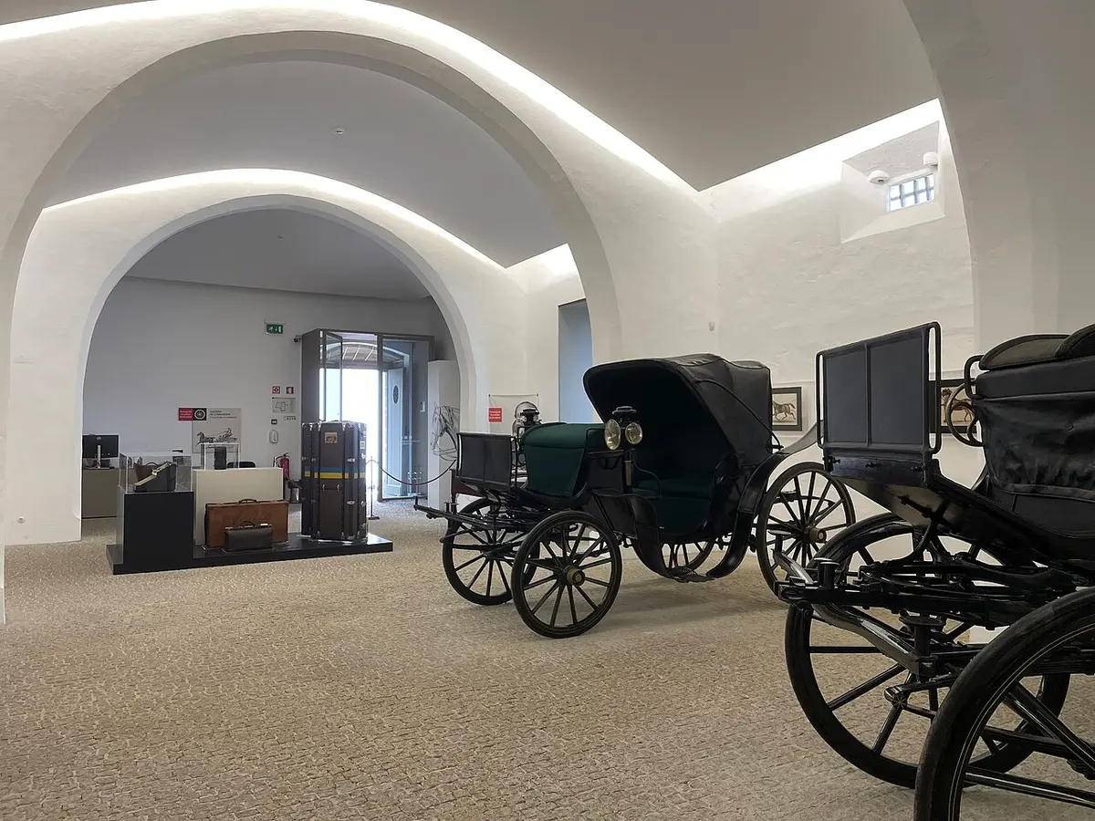 Sala abobadada branca com carruagens antigas e malas de viagem em exposição.