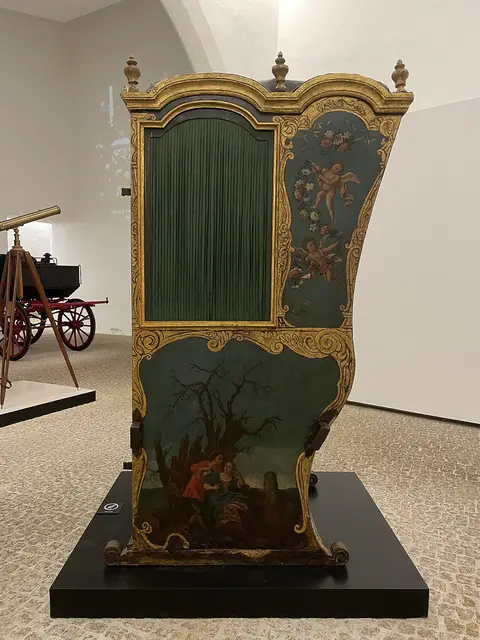 Cadeirinha de liteira antiga, pintada e dourada, exposta numa sala de museu de carruagens