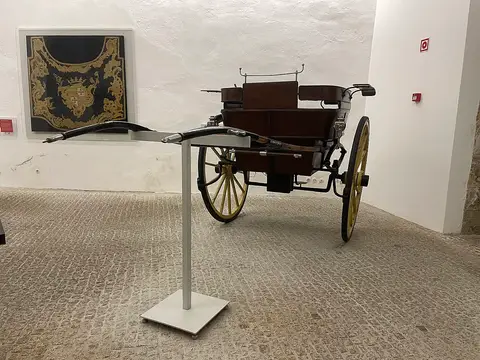 Carruagem antiga de duas rodas exposta em sala de museu com paredes brancas e chão de calçada