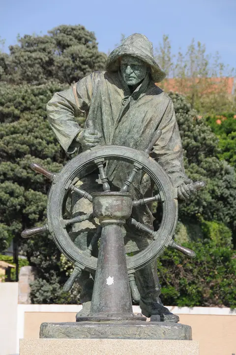 Estátua de bronze do Homem do Leme, pescador ao leme, sobre base de pedra rodeada de vegetação