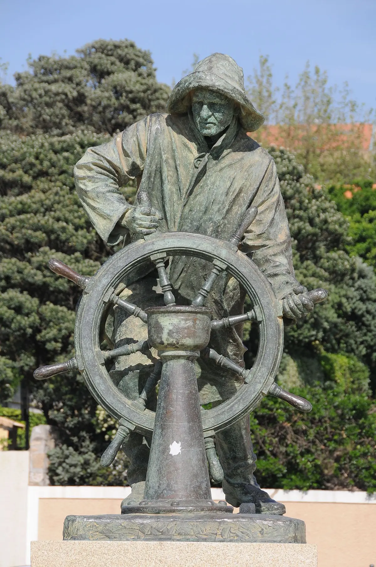 Estátua de bronze do Homem do Leme, pescador ao leme, sobre base de pedra rodeada de vegetação