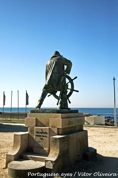Escultura em bronze do Homem do Leme sobre base de granito, junto ao mar, sob céu azul