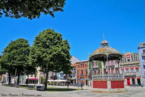 Coreto de ferro forjado numa praça arborizada, rodeado de edifícios coloridos sob céu azul