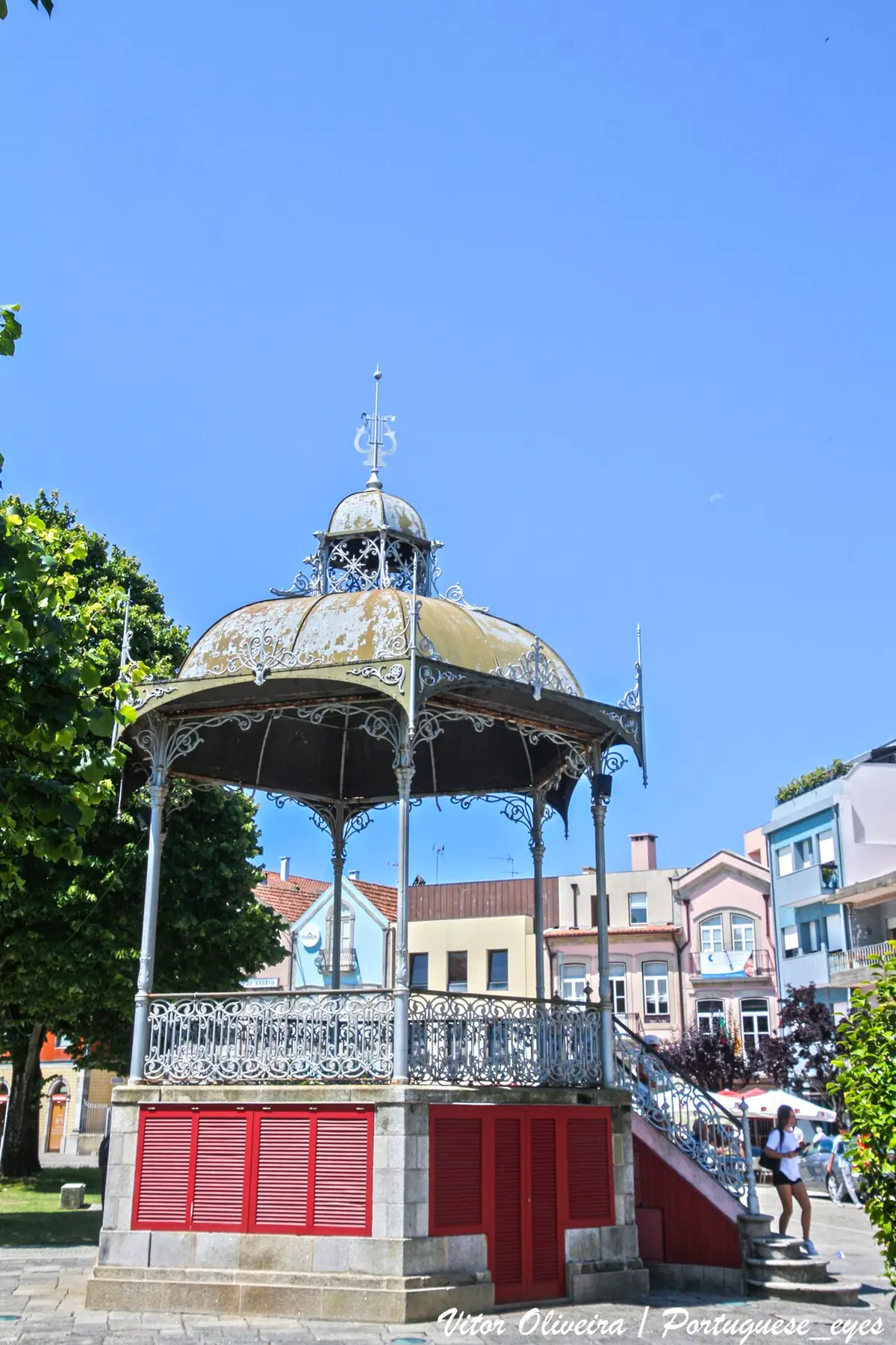 Coreto de ferro forjado com base em pedra e portadas vermelhas, numa praça com edifícios coloridos.