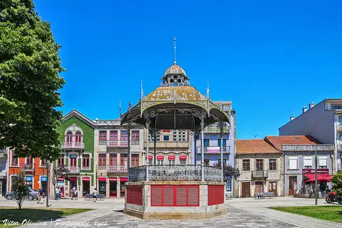 Coreto de ferro forjado com base vermelha numa praça, rodeado de edifícios coloridos e céu azul.