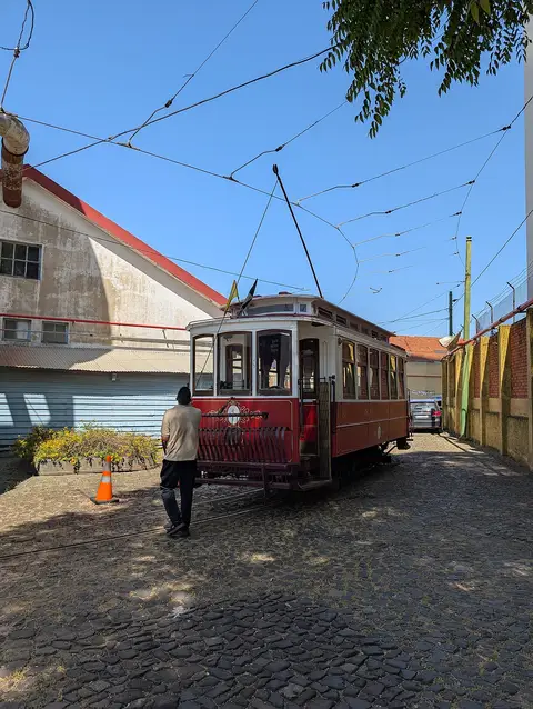 Elétrico antigo vermelho e creme numa via de calçada no Museu da Carris, com pessoa de costas.