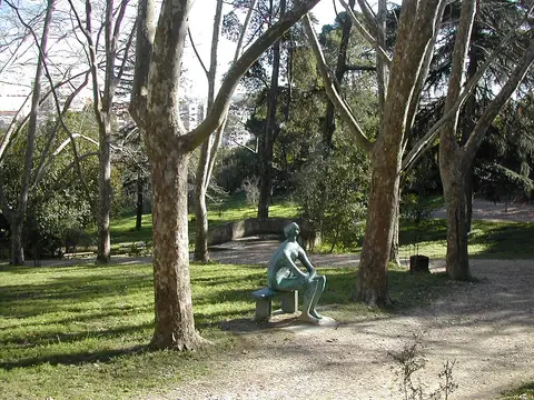 Escultura de bronze de figura sentada num banco, rodeada de árvores e relva no Parque Bensaúde