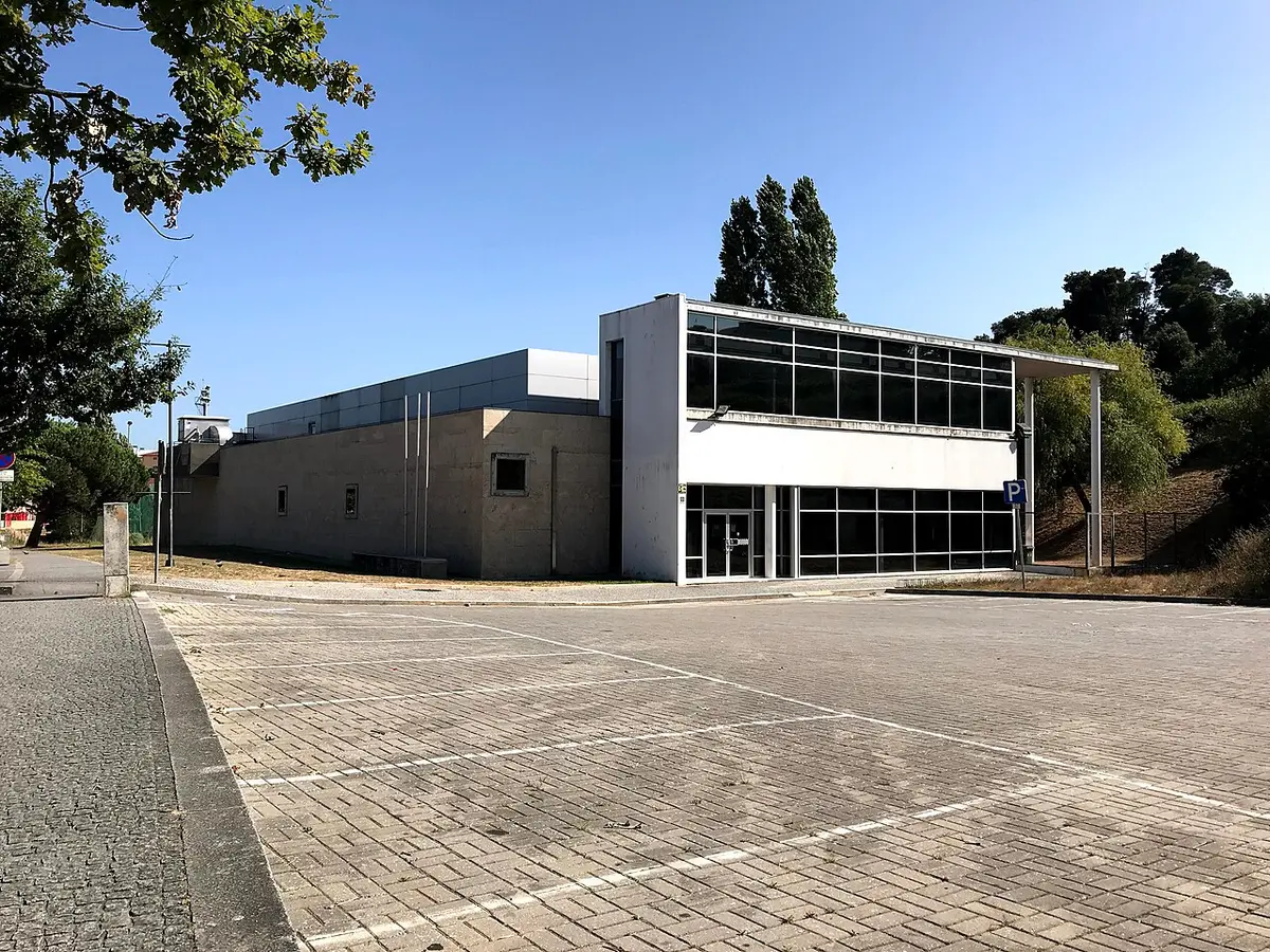 Exterior do edifício da piscina municipal, com fachada envidraçada e parque de estacionamento em frente