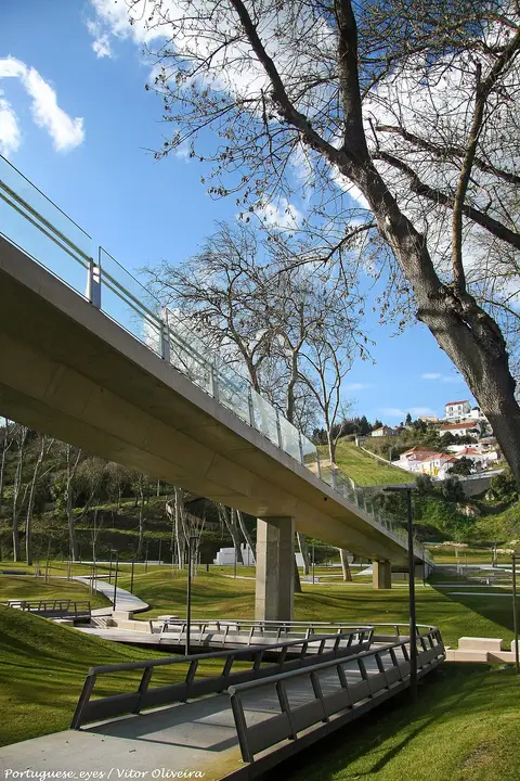 Passadiço elevado em betão e vidro sobre relvados do parque urbano, com árvores e casas ao fundo