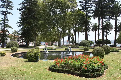 Lago com esculturas e repuxo rodeado de relva, buxos e canteiro de flores vermelhas no Passeio Alegre