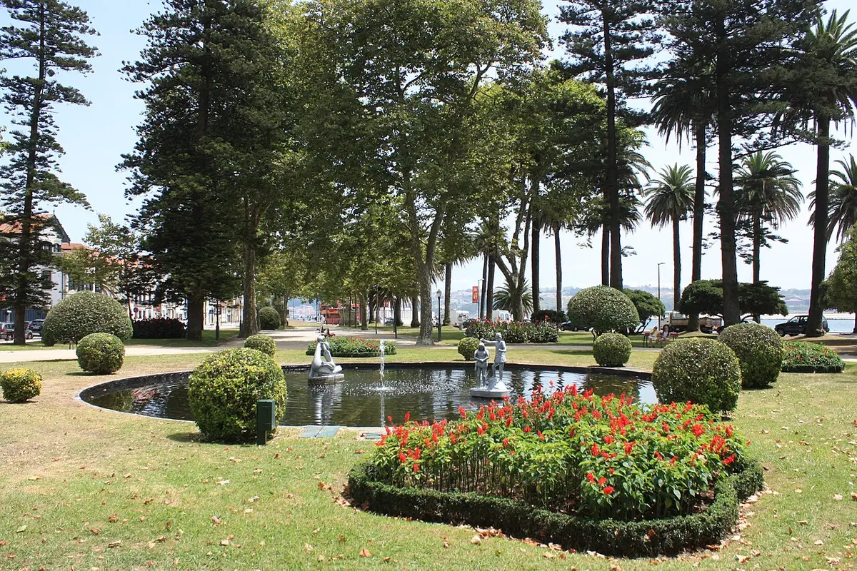 Lago com esculturas e repuxo rodeado de relva, buxos e canteiro de flores vermelhas no Passeio Alegre