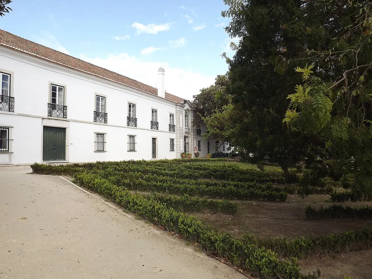 Fachada branca do palácio da Quinta Nossa Senhora da Piedade, com sebes aparadas e árvores no jardim