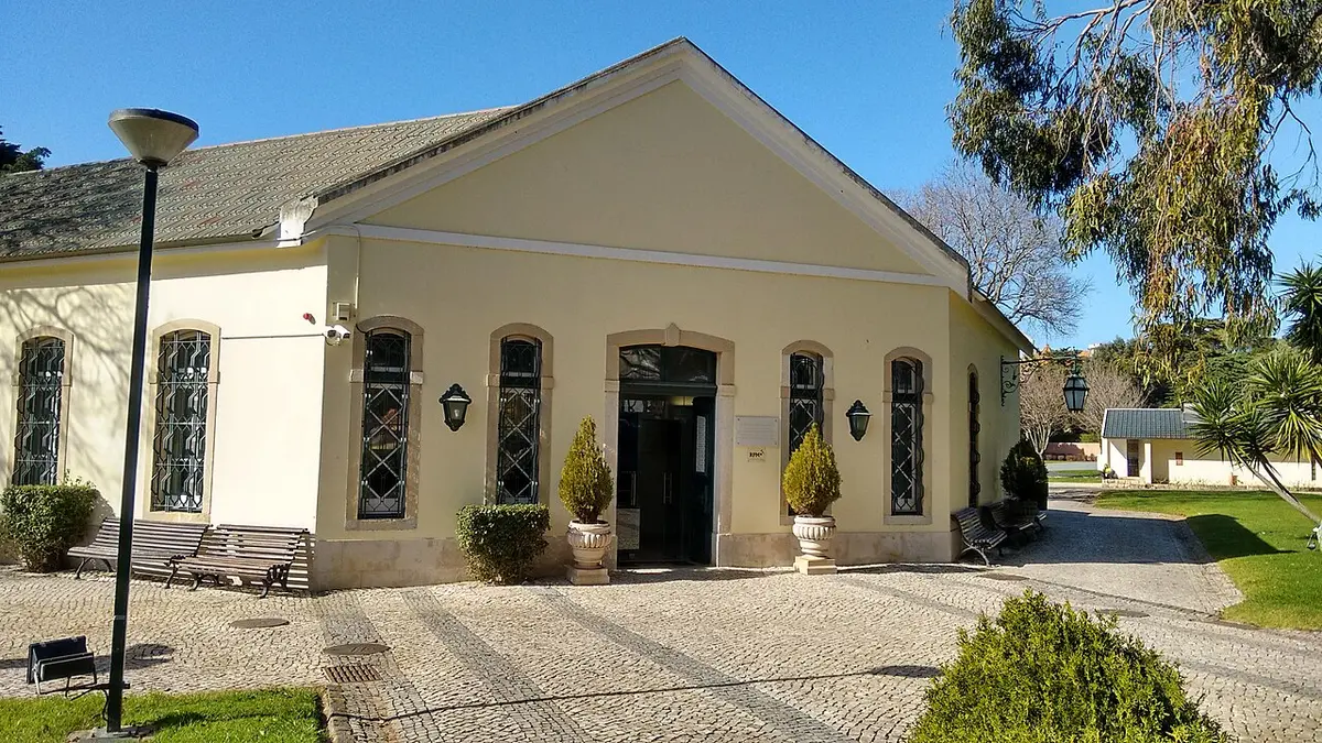 Edifício amarelo com entrada em arco e calçada portuguesa, rodeado de jardim e árvores