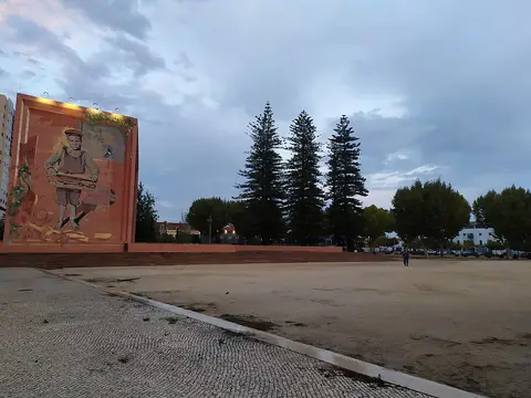 Largo em terra batida no Parque José Afonso, com mural pintado num muro e pinheiros ao fundo