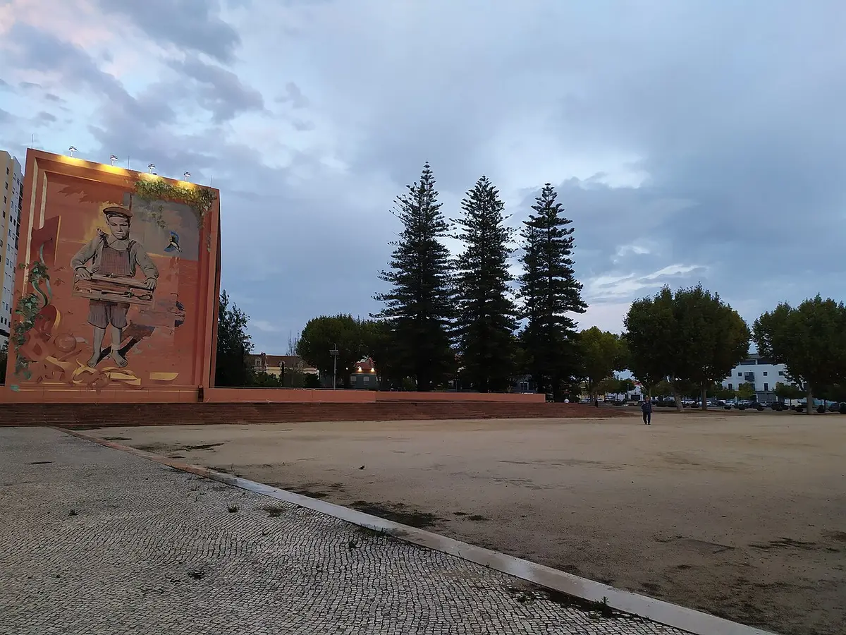 Largo em terra batida no Parque José Afonso, com mural pintado num muro e pinheiros ao fundo