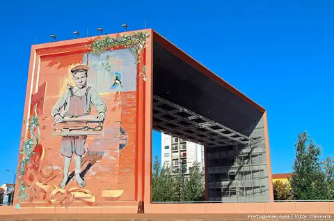 Auditório ao ar livre com mural de um rapaz pintado na fachada cor de terracota, sob céu azul