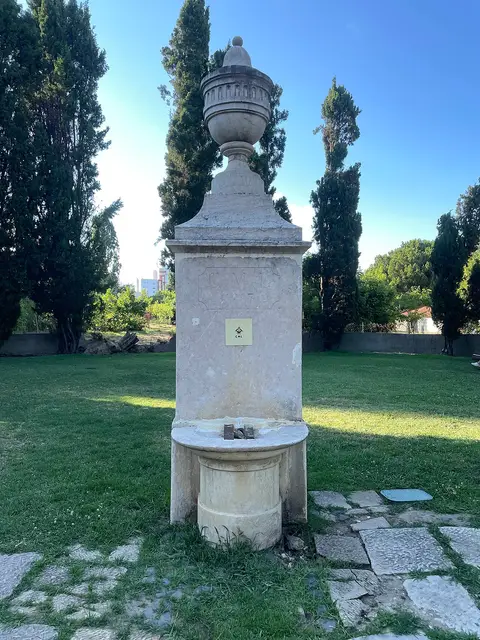 Chafariz de pedra com urna decorativa sobre relva, rodeado de ciprestes no parque urbano