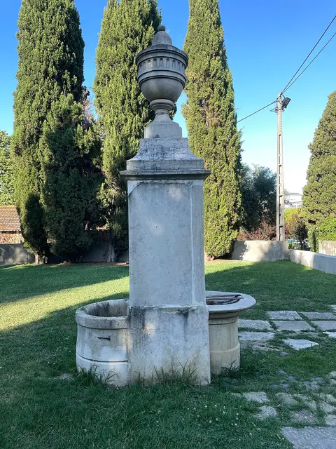 Fonte de pedra com urna decorativa e dois tanques, rodeada de relva e ciprestes no parque