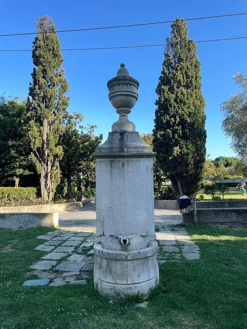 Fonte de pedra encimada por urna decorativa, rodeada de relva e ciprestes no parque da Quinta Conde dos Arcos