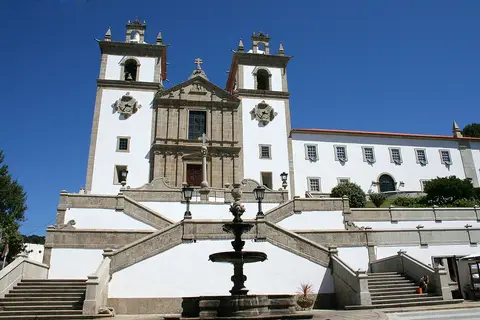 Igreja branca com duas torres sineiras e escadaria com fonte em primeiro plano, sob céu azul.