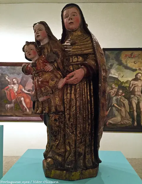 Escultura policromada de Santa Ana com a Virgem e o Menino, exposta no museu, com pinturas ao fundo.