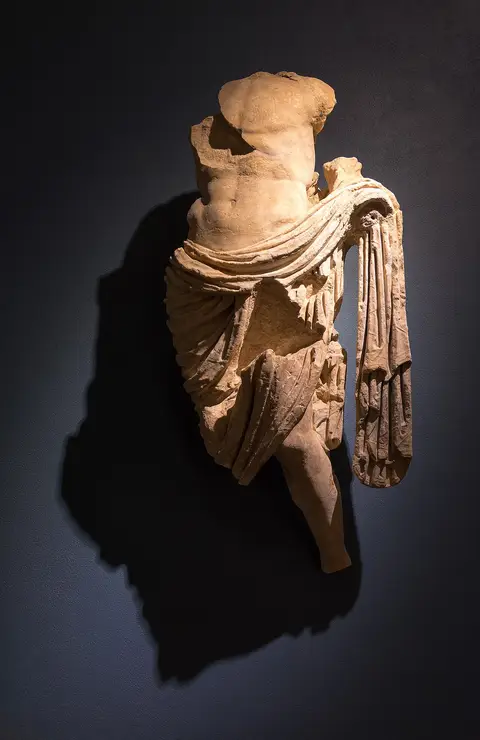 Escultura romana em mármore, torso com túnica drapeada, exposta em parede escura no Museu de Conímbriga