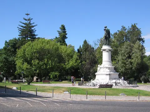 Jardim com relvado, árvores e monumento em pedra com estátua de bronze ao centro